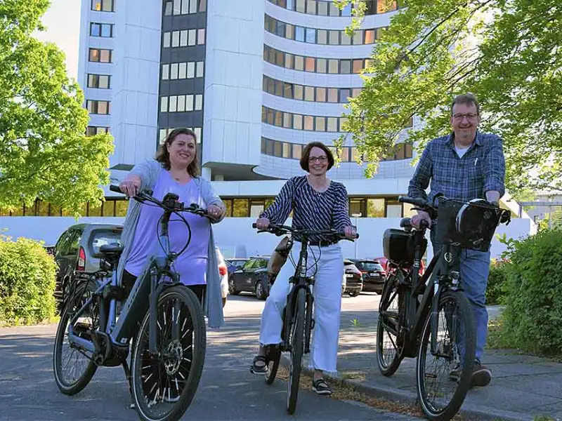 Drei Mitarbeiter:innen, die an einem sonnigen Tag mit dem Fahrrad in Dithmarschen vor einem großen Gebäude stehen